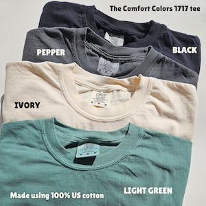 Puede incluir: Cuatro camisetas de diferentes colores: negro, pimienta, marfil y verde claro. Las camisetas est&aacute;n hechas de 100% algod&oacute;n estadounidense y tienen el texto "Comfort Colors 1717 tee" en la parte superior.