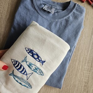 May include: Two t-shirts, one blue and one white. The white t-shirt has an embroidered design of four blue fish.