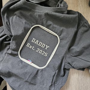 May include: A gray t-shirt with an embroidered design that says "Daddy Est. 2025" in a white embroidery hoop.