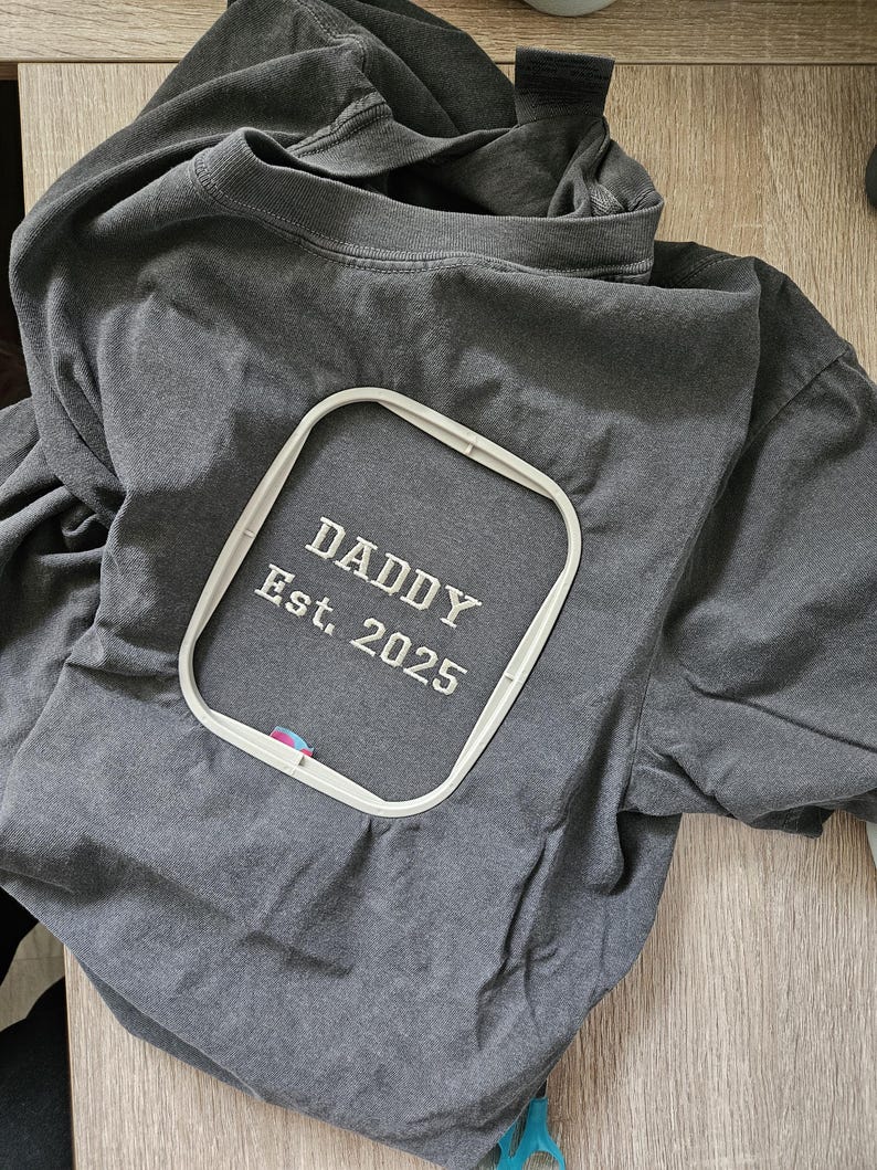 May include: A gray t-shirt with an embroidered design that says "Daddy Est. 2025" in a white embroidery hoop.