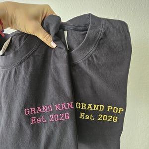 May include: Two dark gray t-shirts with embroidered text. One shirt reads "GRAND NAN Est. 2026" in pink, and the other reads "GRAND POP Est. 2026" in yellow. The shirts are held up against a light background.