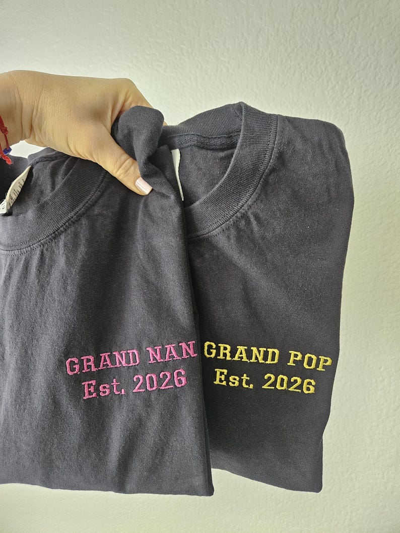 May include: Two dark gray t-shirts with embroidered text. One shirt reads "GRAND NAN Est. 2026" in pink, and the other reads "GRAND POP Est. 2026" in yellow. The shirts are held up against a light background.