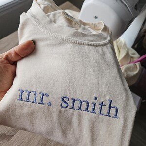May include: Cream-colored t-shirt with the embroidered text "mr. smith" in blue thread. The shirt is folded and held in a person's hand. The shirt appears to be made of a soft, comfortable material.