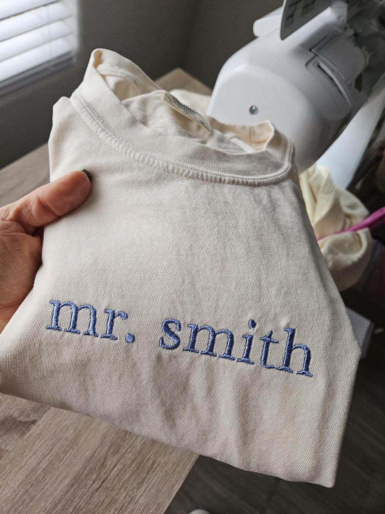 May include: Cream-colored t-shirt with the embroidered text "mr. smith" in blue thread. The shirt is folded and held in a person's hand. The shirt appears to be made of a soft, comfortable material.