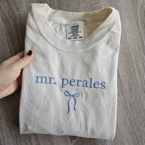 May include: Cream-colored t-shirt with the embroidered text "mr. perales" in blue, along with a blue bow design. The shirt is folded and appears to be made of a soft material. The tag reads "Comfort Colors" and the size is 2XL.