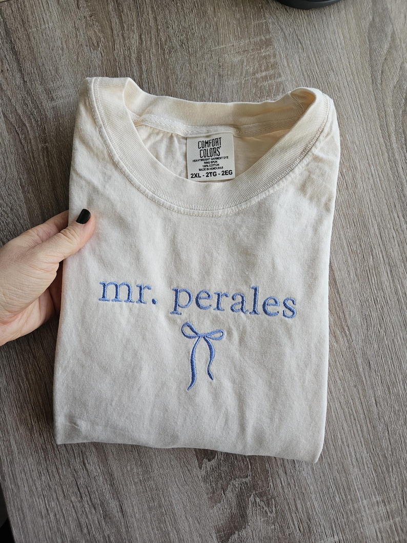 May include: Cream-colored t-shirt with the embroidered text "mr. perales" in blue, along with a blue bow design. The shirt is folded and appears to be made of a soft material. The tag reads "Comfort Colors" and the size is 2XL.