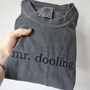 May include: A folded charcoal gray t-shirt with the embroidered text "mr. dooling" in black. The shirt has a crew neck and a tag that reads "Comfort Colors". The shirt is held by a hand.