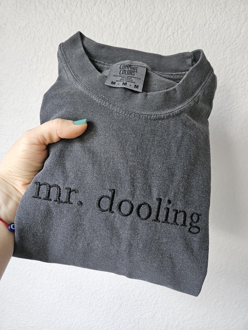 May include: A folded charcoal gray t-shirt with the embroidered text "mr. dooling" in black. The shirt has a crew neck and a tag that reads "Comfort Colors". The shirt is held by a hand.