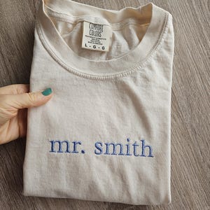 May include: Cream-colored t-shirt with the embroidered text "mr. smith" in blue. The shirt is folded and features a tag with the "Comfort Colors" logo and size information. The shirt is made of heavyweight garment dye cotton.