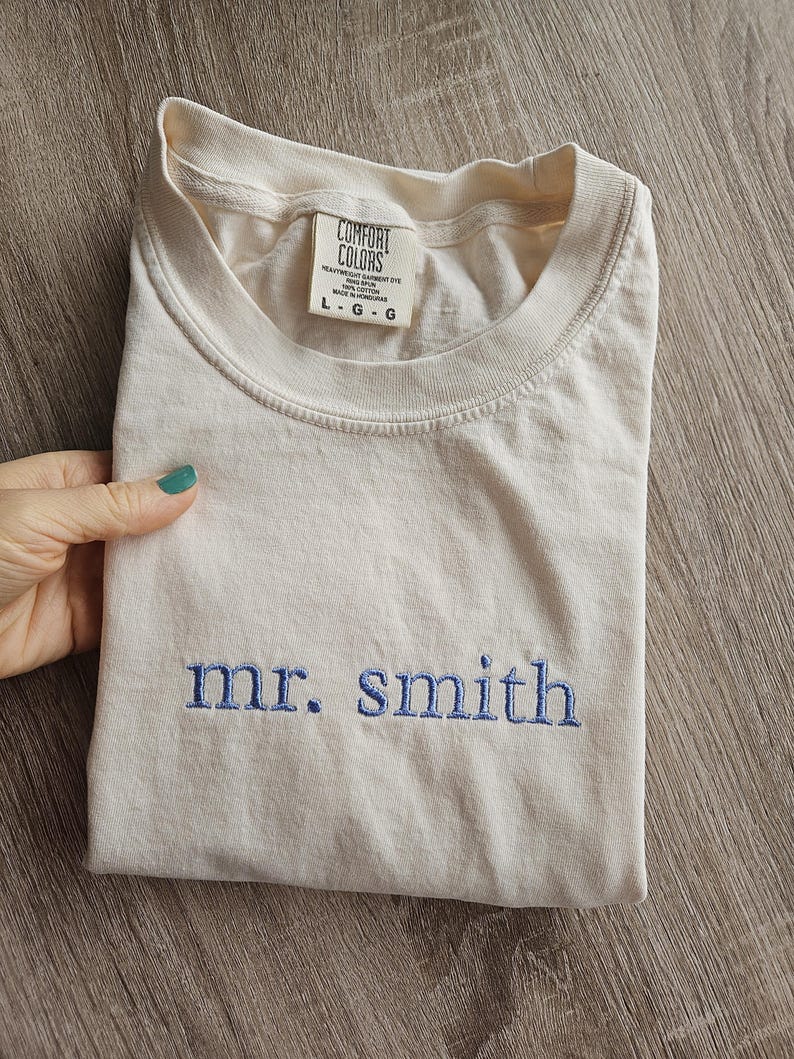 May include: Cream-colored t-shirt with the embroidered text "mr. smith" in blue. The shirt is folded and features a tag with the "Comfort Colors" logo and size information. The shirt is made of heavyweight garment dye cotton.