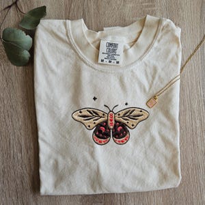 K&ouml;nnte beinhalten: Cremefarbenes Comfort Colors T-Shirt mit einem gestickten Schmetterlingsdesign. Der Schmetterling hat beige Fl&uuml;gel mit schwarzen und roten Details. Eine goldene Halskette mit einem rosa Anh&auml;nger liegt auf dem Shirt. Das Shirt-Etikett lautet "Comfort Colors".