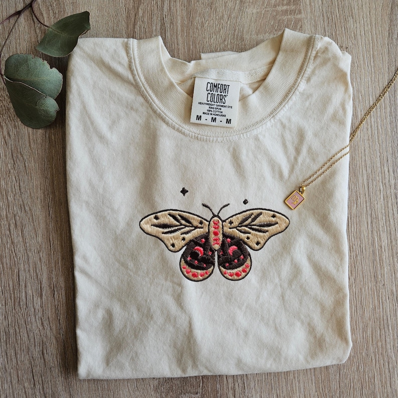 K&ouml;nnte beinhalten: Cremefarbenes Comfort Colors T-Shirt mit einem gestickten Schmetterlingsdesign. Der Schmetterling hat beige Fl&uuml;gel mit schwarzen und roten Details. Eine goldene Halskette mit einem rosa Anh&auml;nger liegt auf dem Shirt. Das Shirt-Etikett lautet "Comfort Colors".