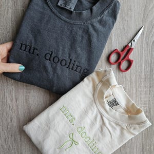 May include: Two folded t-shirts, one charcoal gray and one cream-colored, with embroidered text. The gray shirt says "mr. dooling" and the cream shirt says "mrs. dooling" with a green bow. A pair of red-handled scissors is also visible.