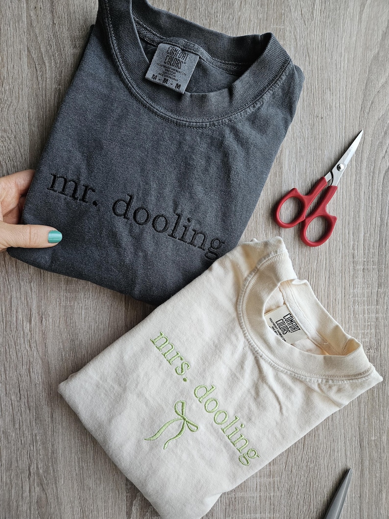 May include: Two folded t-shirts, one charcoal gray and one cream-colored, with embroidered text. The gray shirt says "mr. dooling" and the cream shirt says "mrs. dooling" with a green bow. A pair of red-handled scissors is also visible.