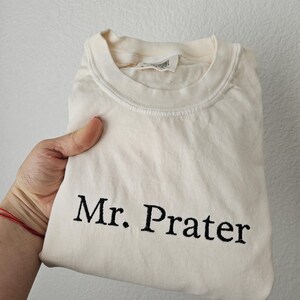 May include: Cream-colored t-shirt with the embroidered text "Mr. Prater" in black. The shirt is folded and held by a hand, showcasing the design. The shirt has a crew neck and a tag.