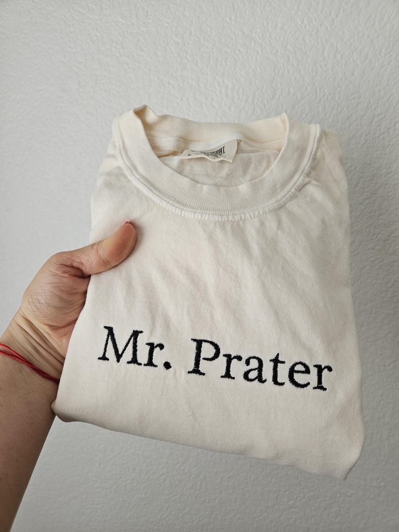 May include: Cream-colored t-shirt with the embroidered text "Mr. Prater" in black. The shirt is folded and held by a hand, showcasing the design. The shirt has a crew neck and a tag.