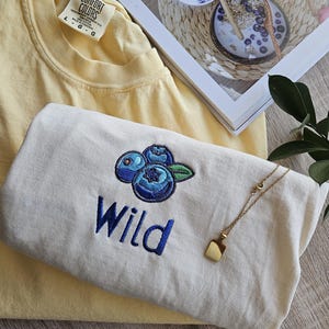 May include: Two t-shirts, one yellow and one white, with the word "Wild" embroidered on the white shirt. The white shirt also features an embroidered design of three blueberries. A gold necklace with a square pendant is on top of the white shirt.