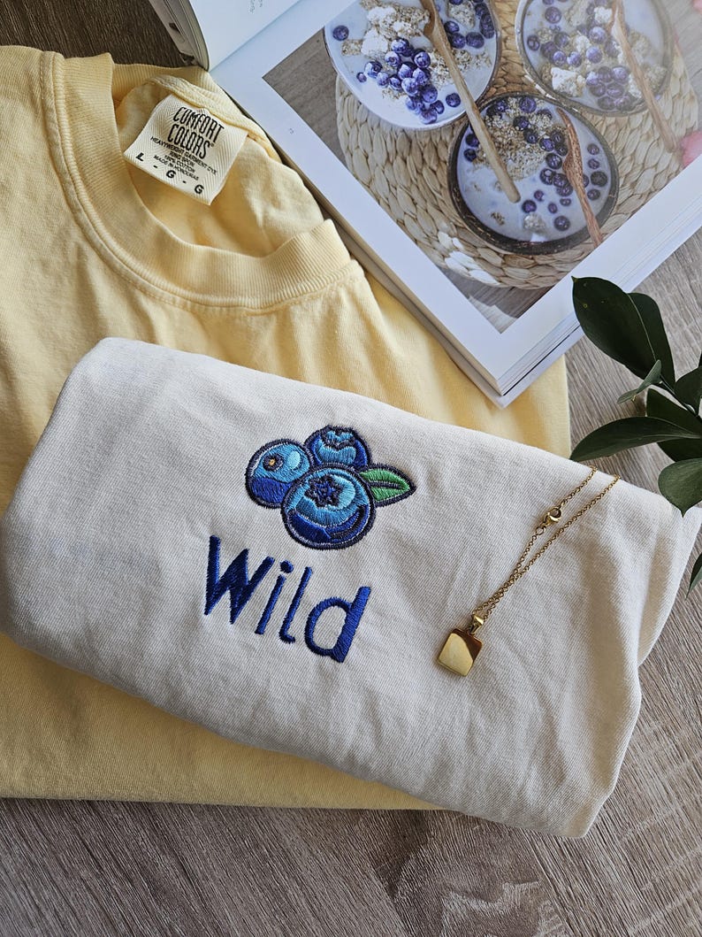 May include: Two t-shirts, one yellow and one white, with the word "Wild" embroidered on the white shirt. The white shirt also features an embroidered design of three blueberries. A gold necklace with a square pendant is on top of the white shirt.