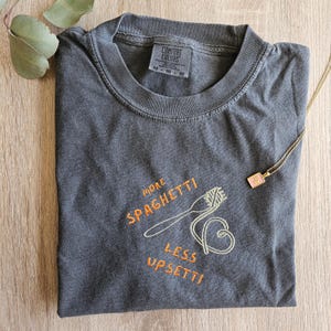 May include: A dark gray t-shirt with the text "More Spaghetti Less Upsetti" embroidered in orange and yellow. The shirt features a small embroidered image of a fork and spaghetti.