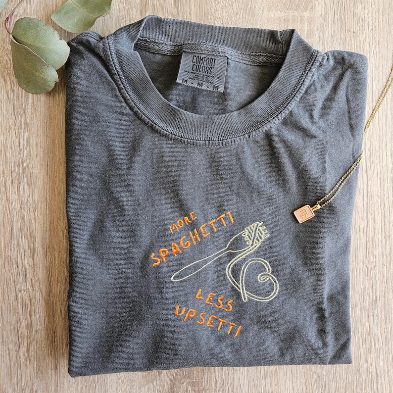 May include: A dark gray t-shirt with the text "More Spaghetti Less Upsetti" embroidered in orange and yellow. The shirt features a small embroidered image of a fork and spaghetti.