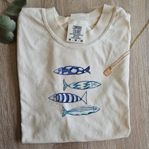 May include: A cream colored t-shirt with a blue and white embroidered design of four fish swimming in a row. The shirt has a tag that reads "COMFORT COLORS" and "HEAVYWEIGHT GARMENT DYE" and "MADE IN HONDURAS".