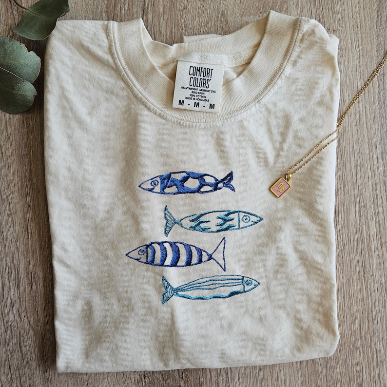 May include: A cream colored t-shirt with a blue and white embroidered design of four fish swimming in a row. The shirt has a tag that reads "COMFORT COLORS" and "HEAVYWEIGHT GARMENT DYE" and "MADE IN HONDURAS".