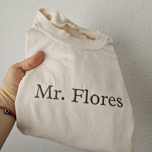 May include: A folded, light beige t-shirt with the embroidered text "Mr. Flores" in brown. The shirt is made of a soft material and has a crew neck.