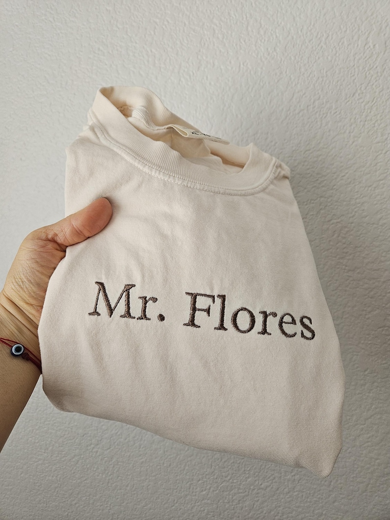 May include: A folded, light beige t-shirt with the embroidered text "Mr. Flores" in brown. The shirt is made of a soft material and has a crew neck.