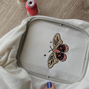K&ouml;nnte beinhalten: Wei&szlig;er Stoff in einem Stickrahmen mit einem detaillierten Schmetterlingsdesign aus beigem, bordeauxrotem und schwarzem Garn. Zwei Spulen rosa Garn liegen daneben. Das Design ist auf dem Stoff zentriert.