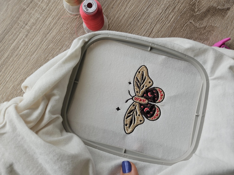 K&ouml;nnte beinhalten: Wei&szlig;er Stoff in einem Stickrahmen mit einem detaillierten Schmetterlingsdesign aus beigem, bordeauxrotem und schwarzem Garn. Zwei Spulen rosa Garn liegen daneben. Das Design ist auf dem Stoff zentriert.