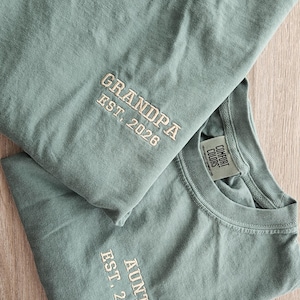 May include: Two sage green t-shirts with embroidered text. One reads "GRANDPA EST. 2026" and the other reads "AUNT EST. 2026". Both shirts have a crew neck and short sleeves. The shirts are made of a soft, comfortable material.