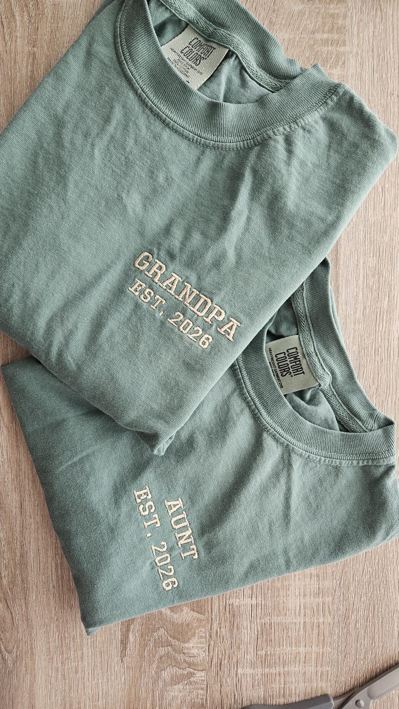 May include: Two sage green t-shirts with embroidered text. One reads "GRANDPA EST. 2026" and the other reads "AUNT EST. 2026". Both shirts have a crew neck and short sleeves. The shirts are made of a soft, comfortable material.