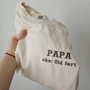 May include: Cream-colored t-shirt with the embroidered text "PAPA aka: Old fart" in brown thread. The shirt has a Comfort Colors tag and is folded.
