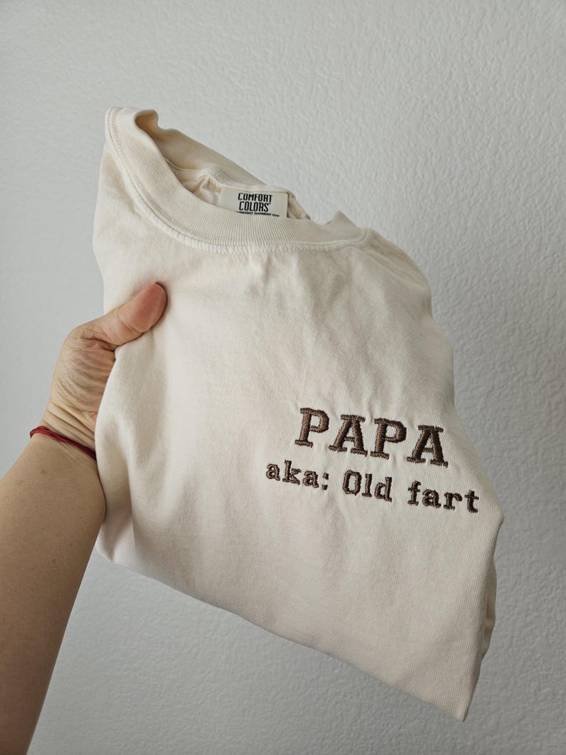 May include: Cream-colored t-shirt with the embroidered text "PAPA aka: Old fart" in brown thread. The shirt has a Comfort Colors tag and is folded.