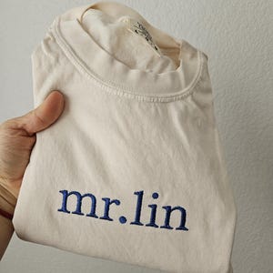 May include: Cream-colored t-shirt with the embroidered text "mr.lin" in navy blue. The shirt is folded, showcasing the text prominently. The fabric appears soft and the overall aesthetic is casual.