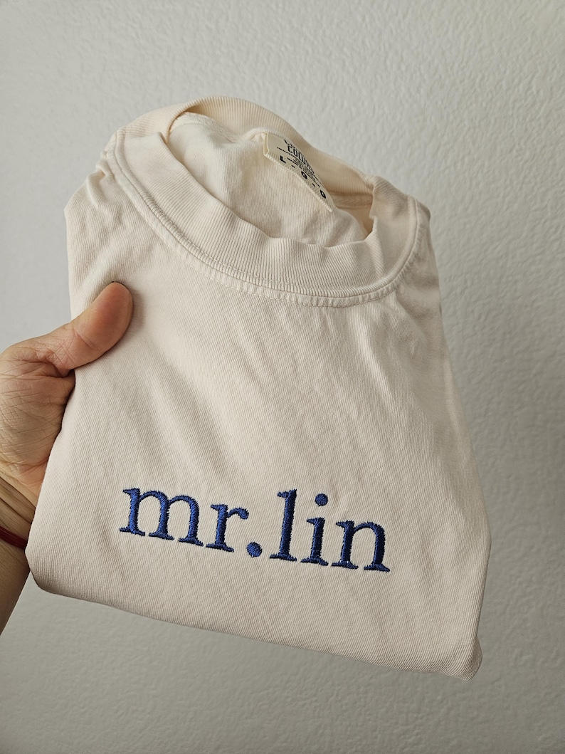 May include: Cream-colored t-shirt with the embroidered text "mr.lin" in navy blue. The shirt is folded, showcasing the text prominently. The fabric appears soft and the overall aesthetic is casual.