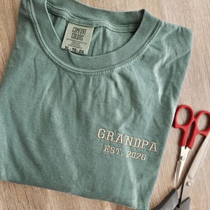 May include: A sage green t-shirt with the word "GRANDPA" embroidered in a light tan thread, along with "EST. 2026". The shirt is folded and next to a pair of scissors.