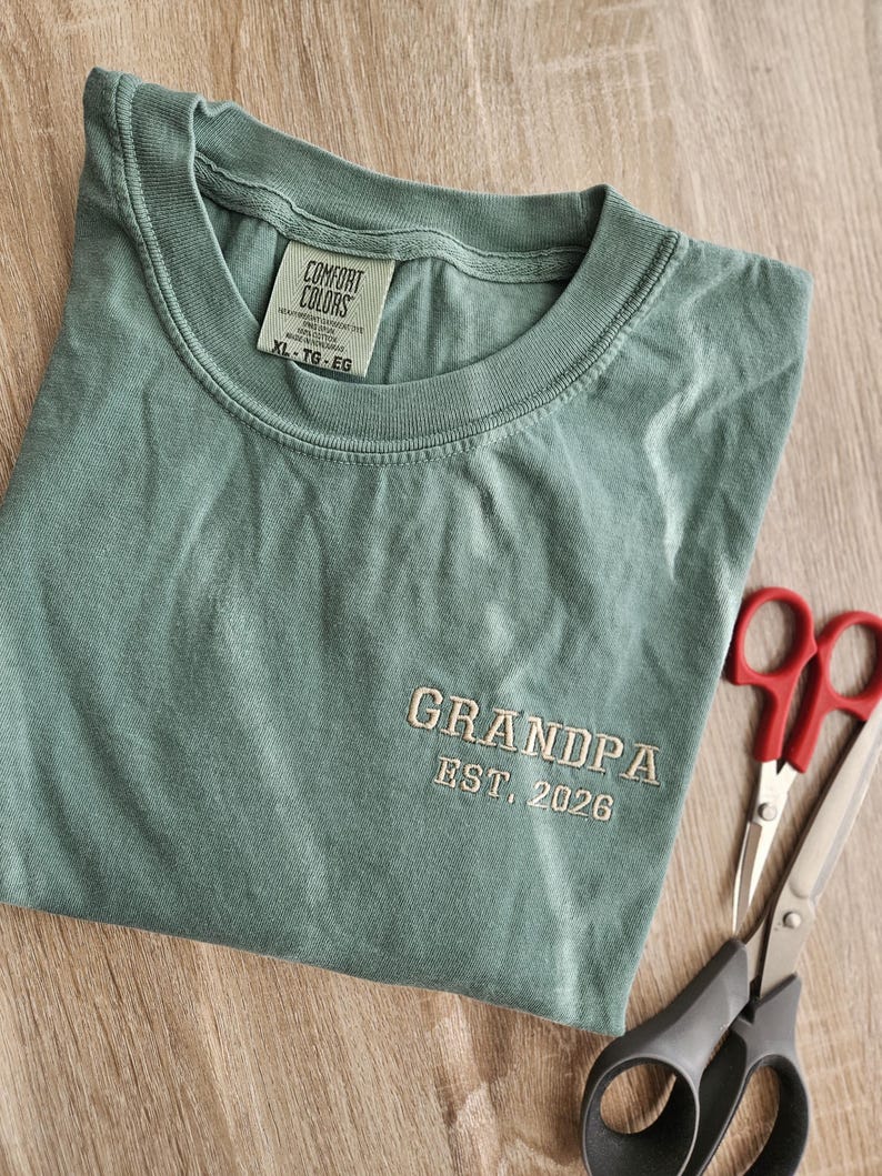 May include: A sage green t-shirt with the word "GRANDPA" embroidered in a light tan thread, along with "EST. 2026". The shirt is folded and next to a pair of scissors.