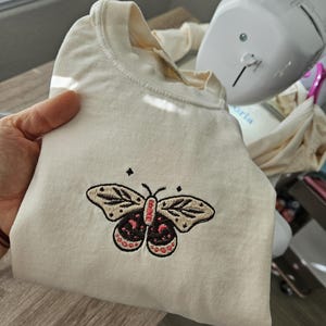 K&ouml;nnte beinhalten: Gefaltetes cremefarbenes T-Shirt mit einem gestickten Schmetterlingsdesign. Der Schmetterling hat beige Fl&uuml;gel mit schwarzen Details und einen rot-schwarzen K&ouml;rper. Das Design befindet sich mittig auf der Vorderseite.