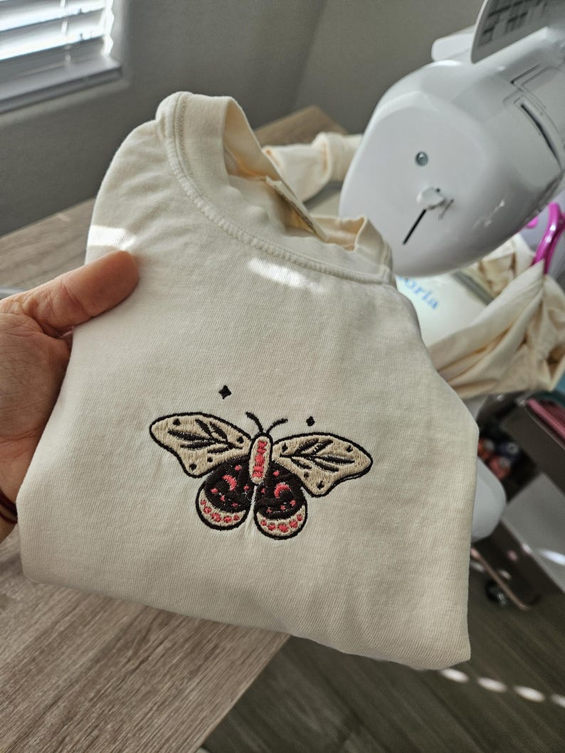 K&ouml;nnte beinhalten: Gefaltetes cremefarbenes T-Shirt mit einem gestickten Schmetterlingsdesign. Der Schmetterling hat beige Fl&uuml;gel mit schwarzen Details und einen rot-schwarzen K&ouml;rper. Das Design befindet sich mittig auf der Vorderseite.