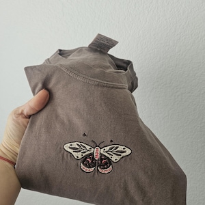 K&ouml;nnte beinhalten: Ein gefaltetes braunes T-Shirt mit einem gestickten Schmetterlingsdesign. Der Schmetterling hat cremefarbene Fl&uuml;gel mit schwarzen und rosa Details. Das Shirt besteht aus einem weichen Material und hat ein Etikett.