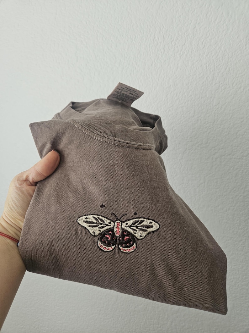 K&ouml;nnte beinhalten: Ein gefaltetes braunes T-Shirt mit einem gestickten Schmetterlingsdesign. Der Schmetterling hat cremefarbene Fl&uuml;gel mit schwarzen und rosa Details. Das Shirt besteht aus einem weichen Material und hat ein Etikett.