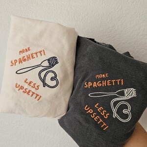 May include: Two t-shirts with embroidered designs. One is beige with the text "More Spaghetti Less Upsetti" and a fork and spaghetti. The other is dark gray with the same text and design.