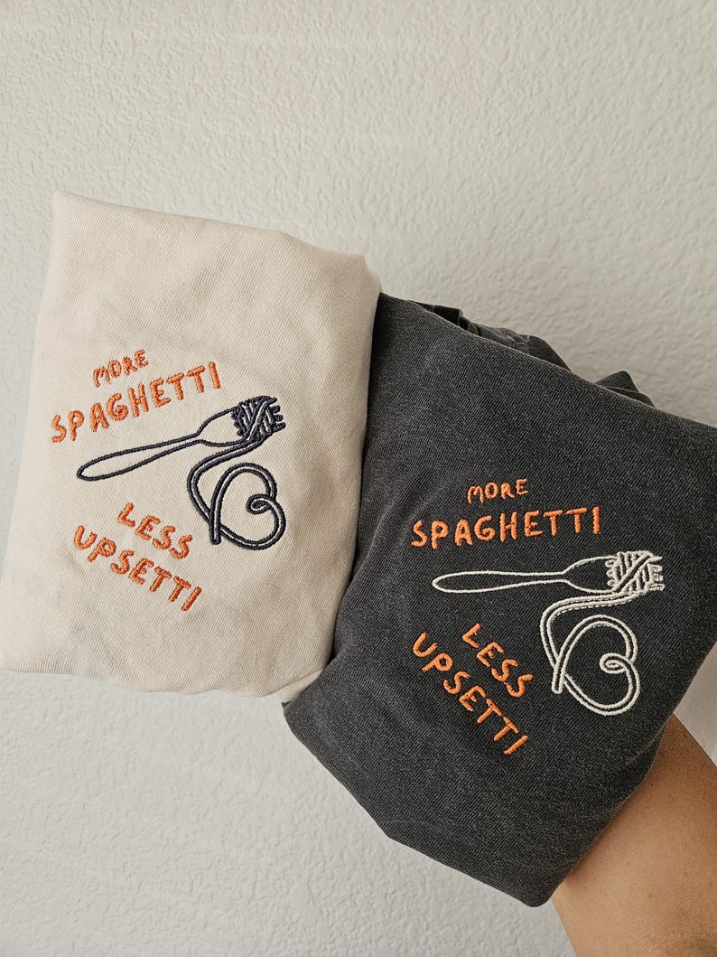 May include: Two t-shirts with embroidered designs. One is beige with the text "More Spaghetti Less Upsetti" and a fork and spaghetti. The other is dark gray with the same text and design.