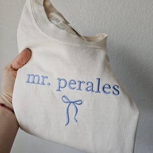 May include: Cream-colored t-shirt with the embroidered text "mr. perales" in blue, along with a blue bow design. The shirt is folded, showcasing the design on the front.