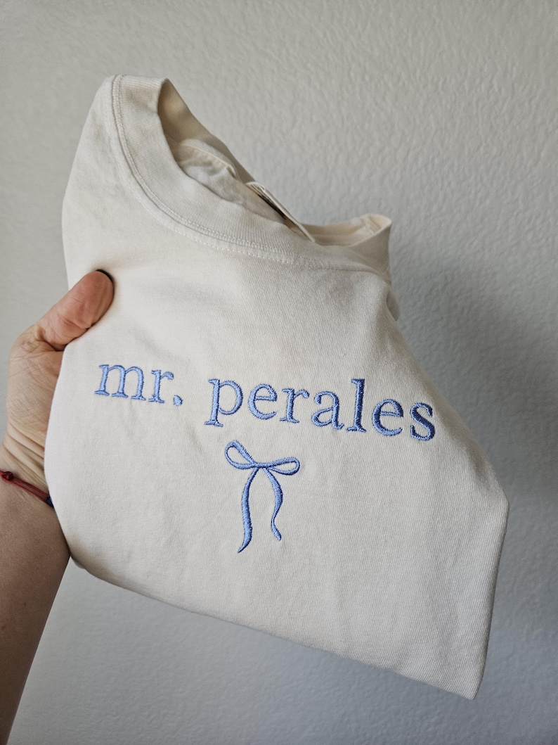 May include: Cream-colored t-shirt with the embroidered text "mr. perales" in blue, along with a blue bow design. The shirt is folded, showcasing the design on the front.