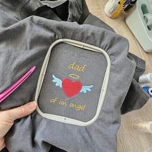 Puede incluir: Una camiseta gris con un dise&ntilde;o bordado de un coraz&oacute;n con alas y un halo sobre &eacute;l. El texto "dad of an angel" est&aacute; bordado debajo del coraz&oacute;n.
