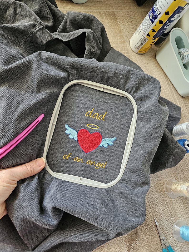 Puede incluir: Una camiseta gris con un dise&ntilde;o bordado de un coraz&oacute;n con alas y un halo sobre &eacute;l. El texto "dad of an angel" est&aacute; bordado debajo del coraz&oacute;n.