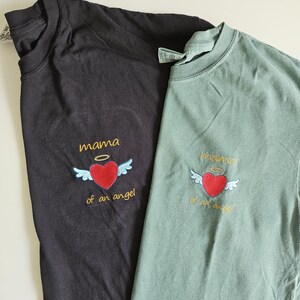 Puede incluir: Dos camisetas con el texto "mama of an angel" bordado en la parte delantera. Las camisetas son negra y verde.