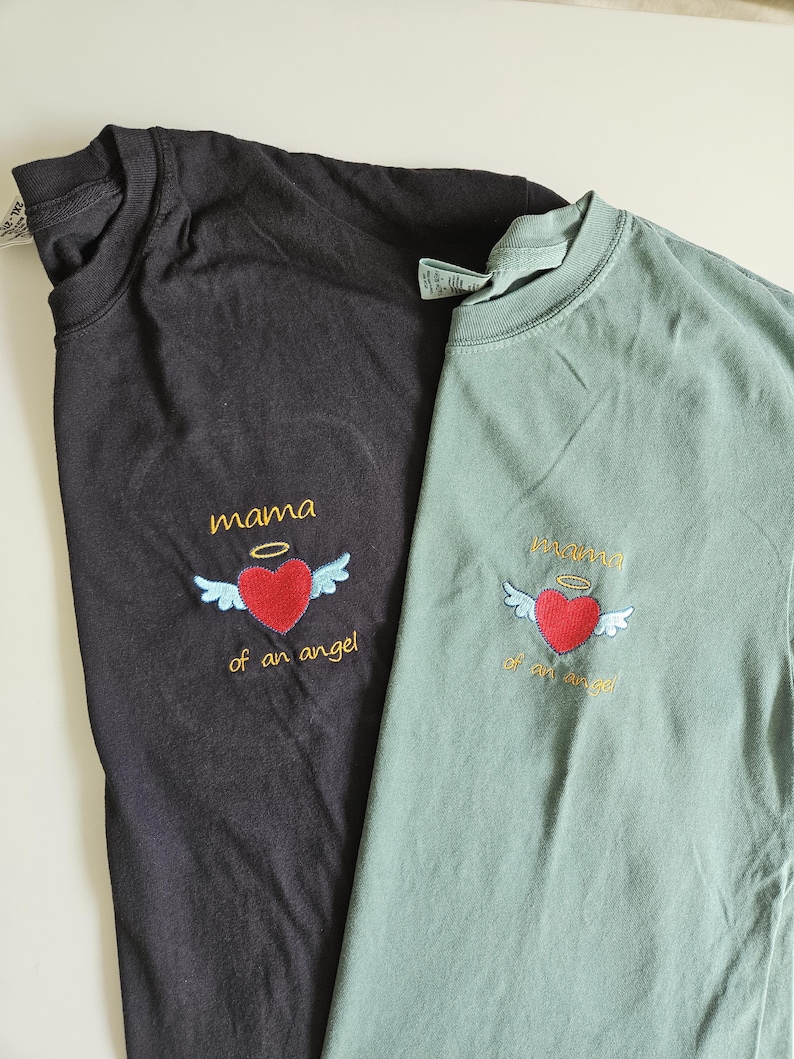 Puede incluir: Dos camisetas con el texto "mama of an angel" bordado en la parte delantera. Las camisetas son negra y verde.